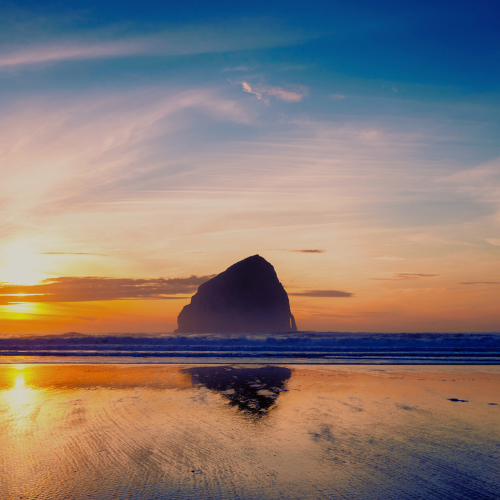 A sea stack silhouetted at sunset on the Oregon coast, reflected in the wet sand